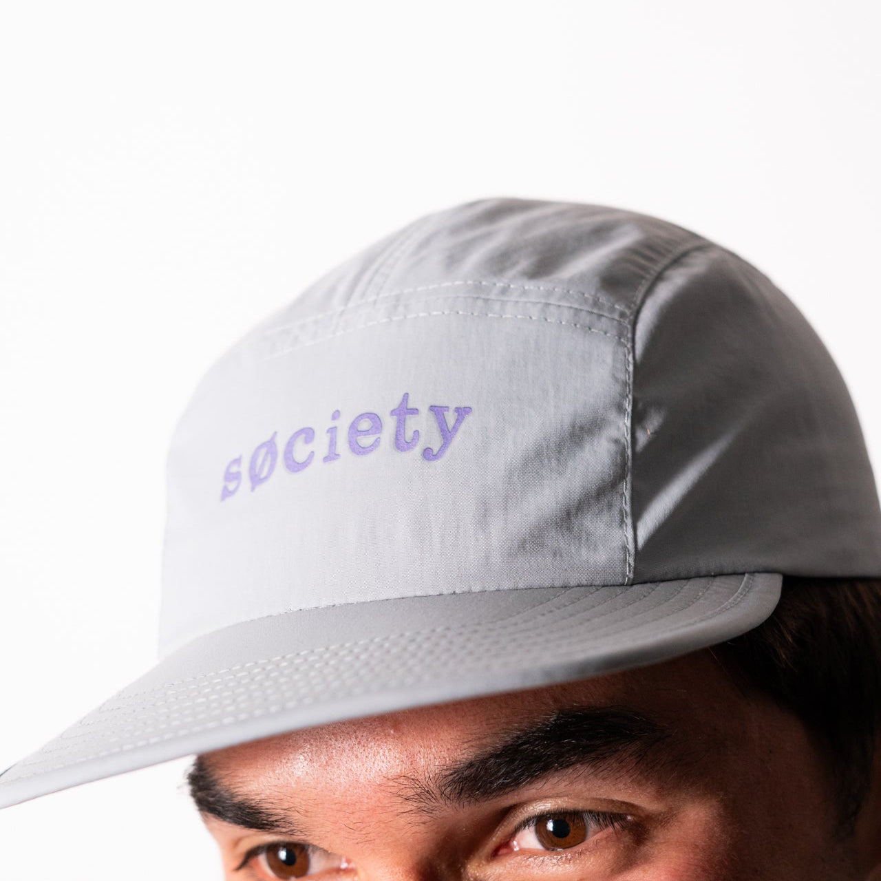 A close-up of a man wearing a light gray 5-panel cap with the word 'society' embroidered in purple on the front, against a plain white background.