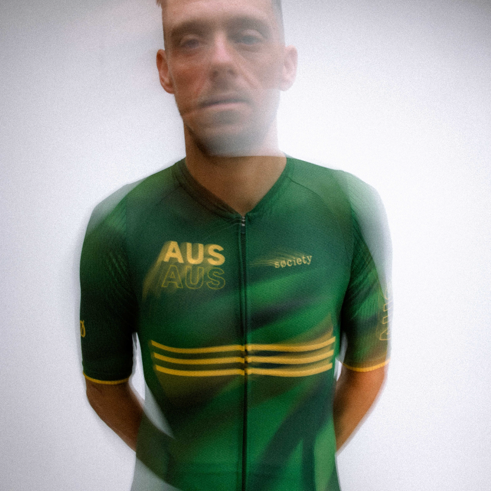 A man wearing a green cycling jersey with yellow stripes and 'AUS' printed on the chest, standing against a light gray background.
