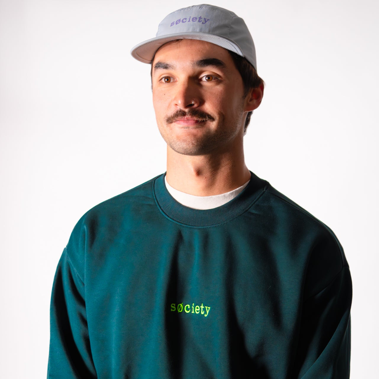 Man wearing a green sweatshirt with 'society' text and a white cap on a white background