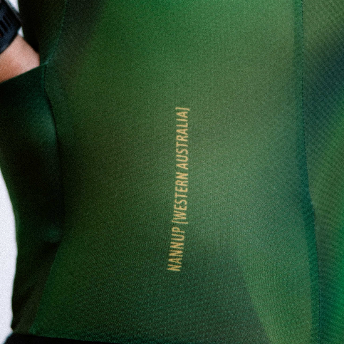 Close-up of a person wearing a green athletic jersey with yellow stripes and vertical 'MANUPOO WESTERN AUSTRALIA' text, paired with black leggings against a plain light background.
