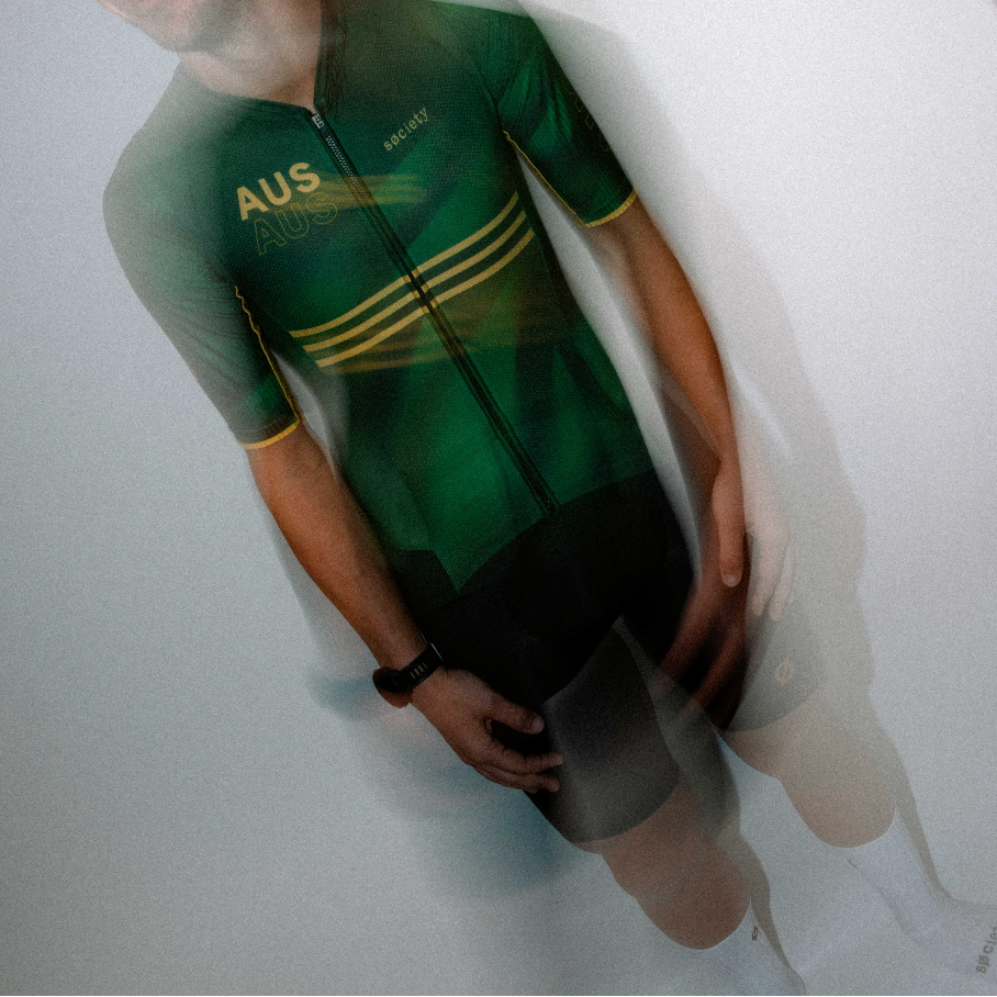 A man wearing a green and black cycling jersey with gold stripes and 'AUS' printed on the chest, posed against a light gray background with a motion-blur overlay.